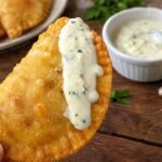 Pastelito venezolano frito con salsa de ajo venezolana cremosa por encima y un recipiente con más salsa al fondo