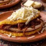 Cachapa venezolana de jojoto doblada sobre un plato de barro, rellena de queso de mano y carne mechada, coronada con queso blanco rallado